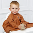 A young child sitting in a white high chair wearing a brown coverall bib with rainbow details, holding a silicone open cup. The background is a clean white panelled wall.