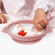 Baby in a pink floral bib using a pink silicone suction bowl with yogurt and strawberries, holding a pink spoon.