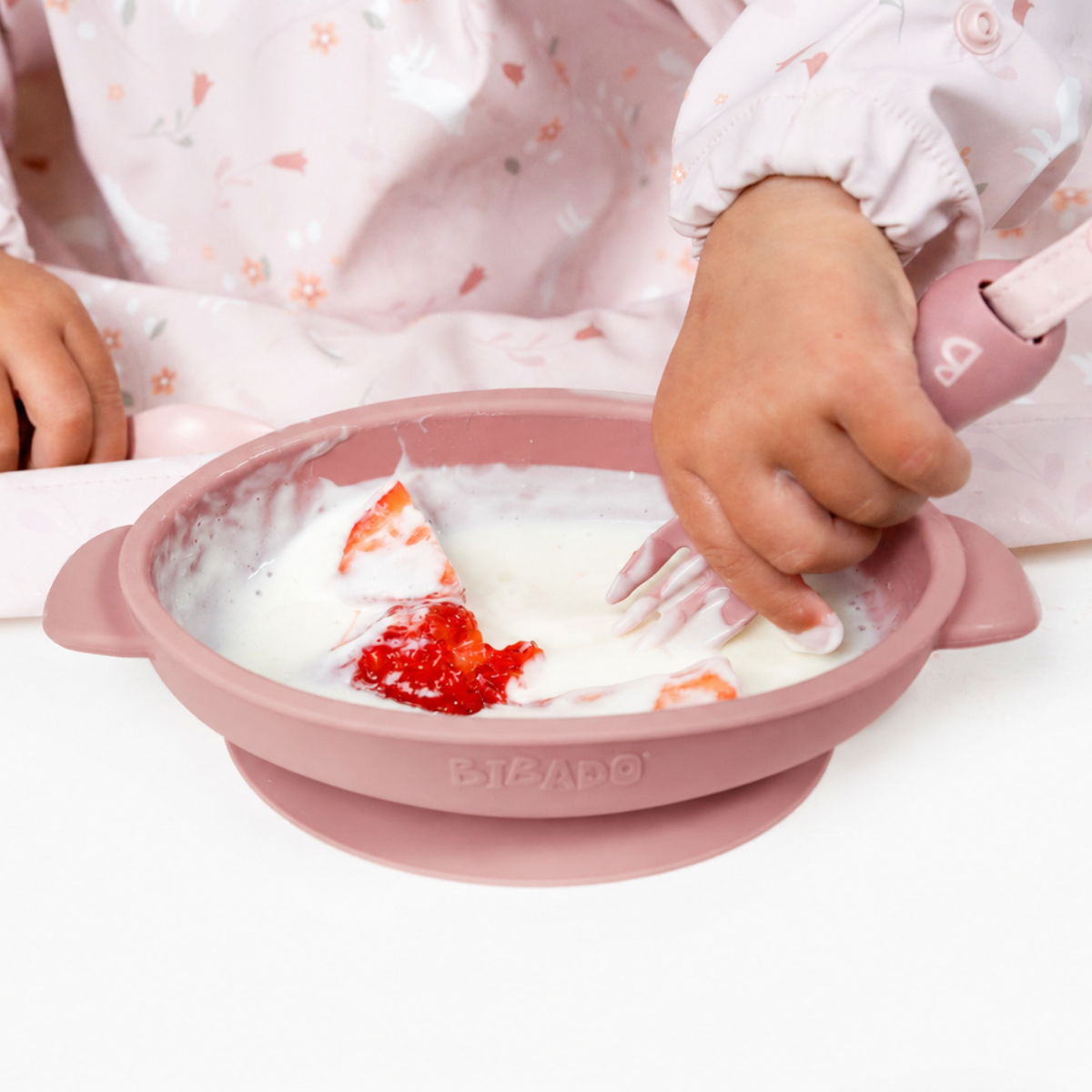 Baby in a pink floral bib using a pink silicone suction bowl with yogurt and strawberries, holding a pink spoon.