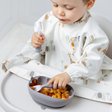 A baby wearing a white long-sleeve bib with a safari animal print sits in a high chair, using a blue silicone suction bowl and matching spoon to eat pasta.