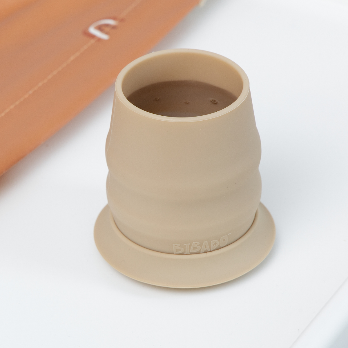 A beige silicone open cup with a matching base ring, shown on a white surface with part of an orange-brown sleeve visible in the background. - Product image