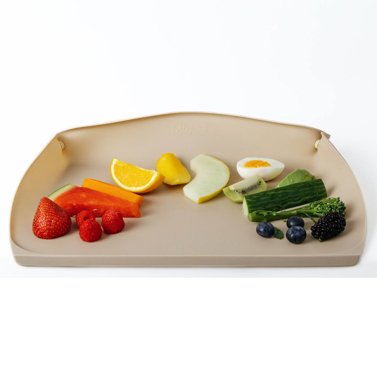 A beige silicone baby feeding mat with optional sides pulled up, shown on a clean white background and filled with colourful fruits, vegetables, and a small egg half. - Product image