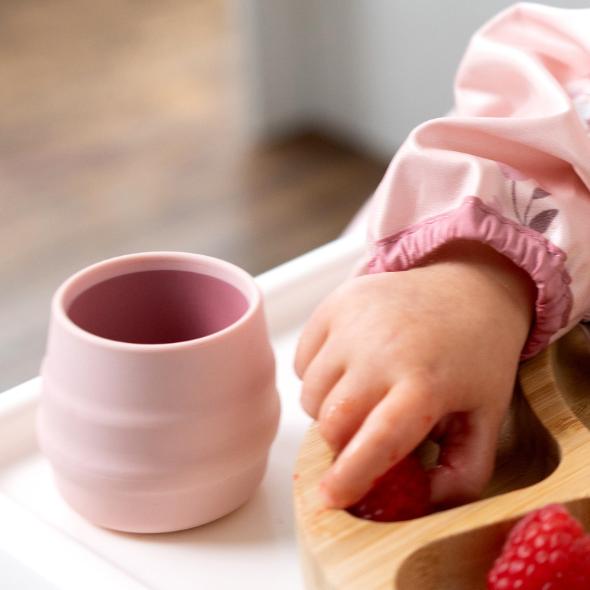 Pink silicone Sippit training cup next to a baby's hand reaching for a strawberry on a bamboo plate. - Product image