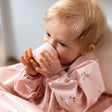 Baby drinking from a small, blush pink silicone open training cup, wearing a matching floral bib.