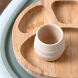 Bibado Sippit™ silicone open training cup in a light beige color, ribbed, on a wooden divided plate.