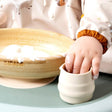 Baby's hand in a small, ribbed, beige silicone Sippit™ open training cup next to a wooden bowl.
