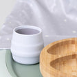 White silicone Sippit™ open training cup with ribbed design next to a wooden plate.