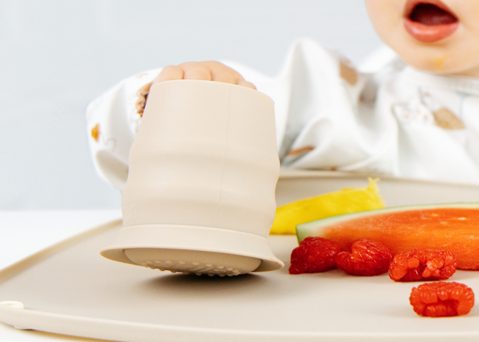 A baby in a white bib reaches for a beige silicone cup with a stabilising base on a beige high chair tray, with food pieces nearby. - product info image