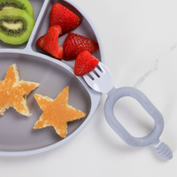 Gray silicone Sporkit™ baby spork and dipper with a strawberry on its tines, next to a divided plate with food.