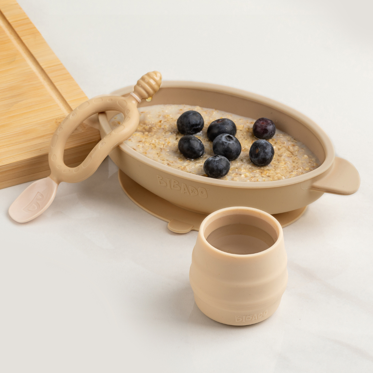 Beige silicone suction bowl with oatmeal and blueberries, matching cup and spoon. - Product image