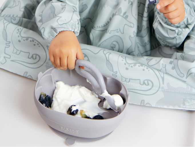 A baby wearing a light blue dinosaur-print bib sits in a high chair, reaching into a gray silicone bowl with a spoon while eating yogurt and blueberries. - image