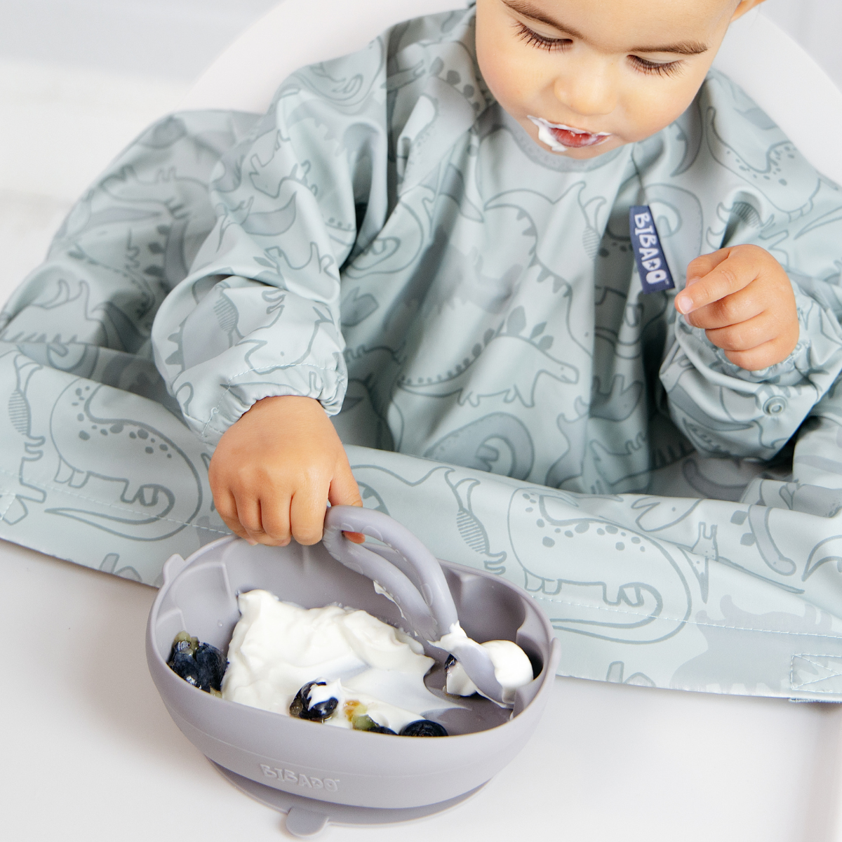A baby wearing a light blue dinosaur-print bib sits in a high chair, reaching into a matching silicone bowl with a spoon while eating yogurt and blueberries. - Product image