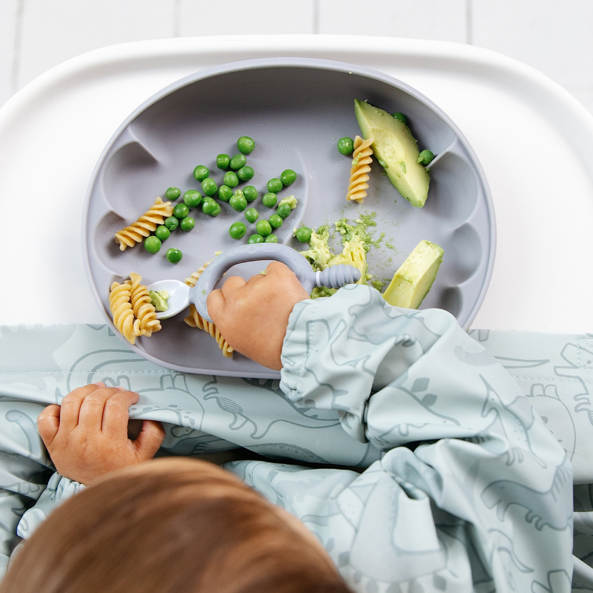 A baby wearing a light blue dinosaur-print bib sits in a high chair, reaching for food on a blue scalloped silicone plate. The plate contains pasta, green peas, and slices of avocado. - Product image