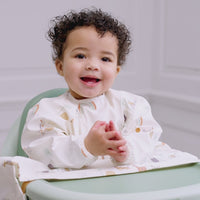 Baby in a long-sleeved, sage green Bibado Coverall Weaning Bib with dinosaur print, eating messy food. - Product image