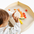 A baby sits in front of a beige feeding mat with the sides raised, spilling a small cup full of water onto the mat. The mat is styled with fruit pieces (watermelon, mango, strawberries) and a small beige cup, set against a clean white background.
