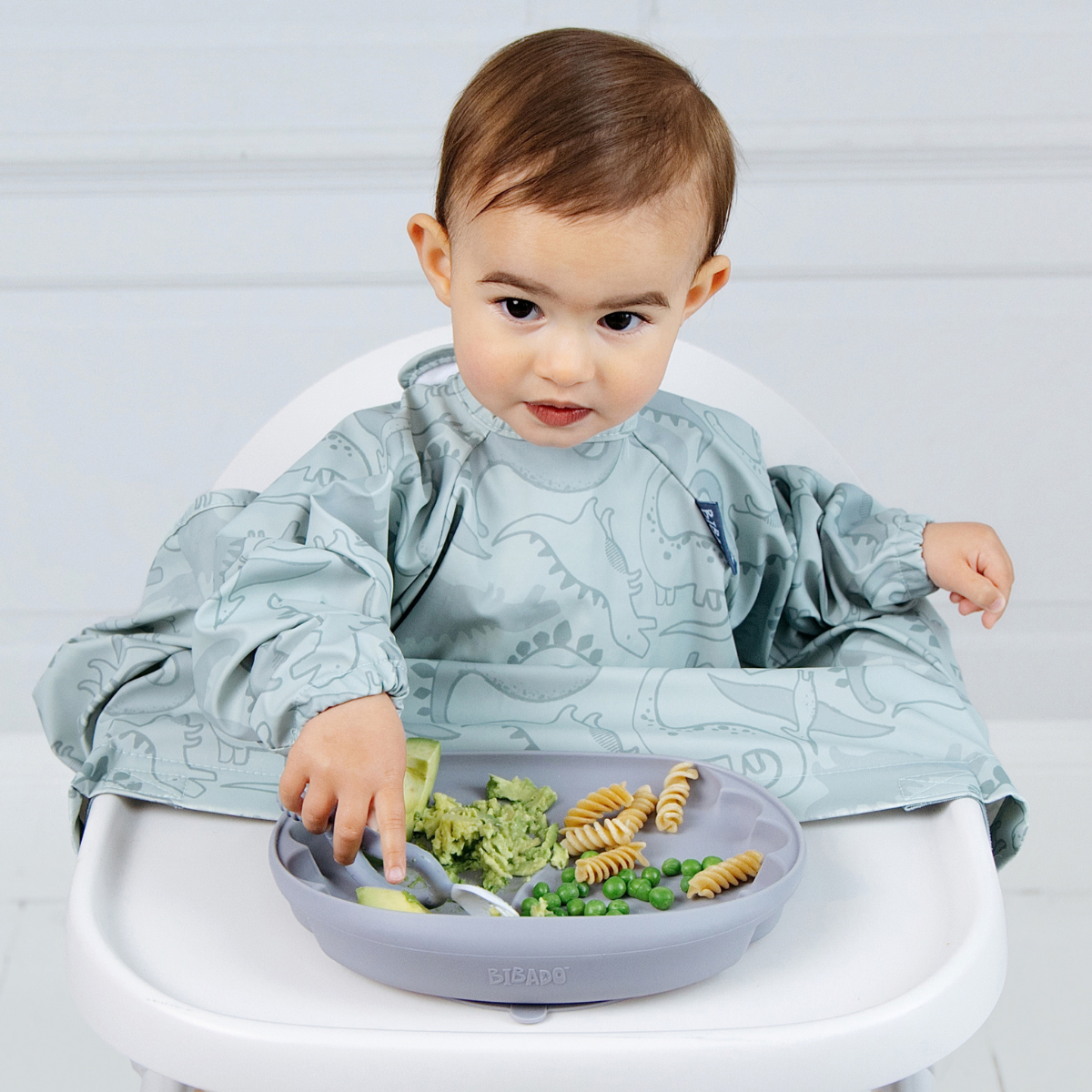 A baby wearing a light blue dinosaur-print bib sits in a white high chair, reaching for food on a matching blue plate. - Product image
