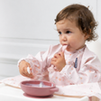 Baby in a high chair wearing a pink floral bib, eating from a pink silicone suction bowl with matching cutlery.