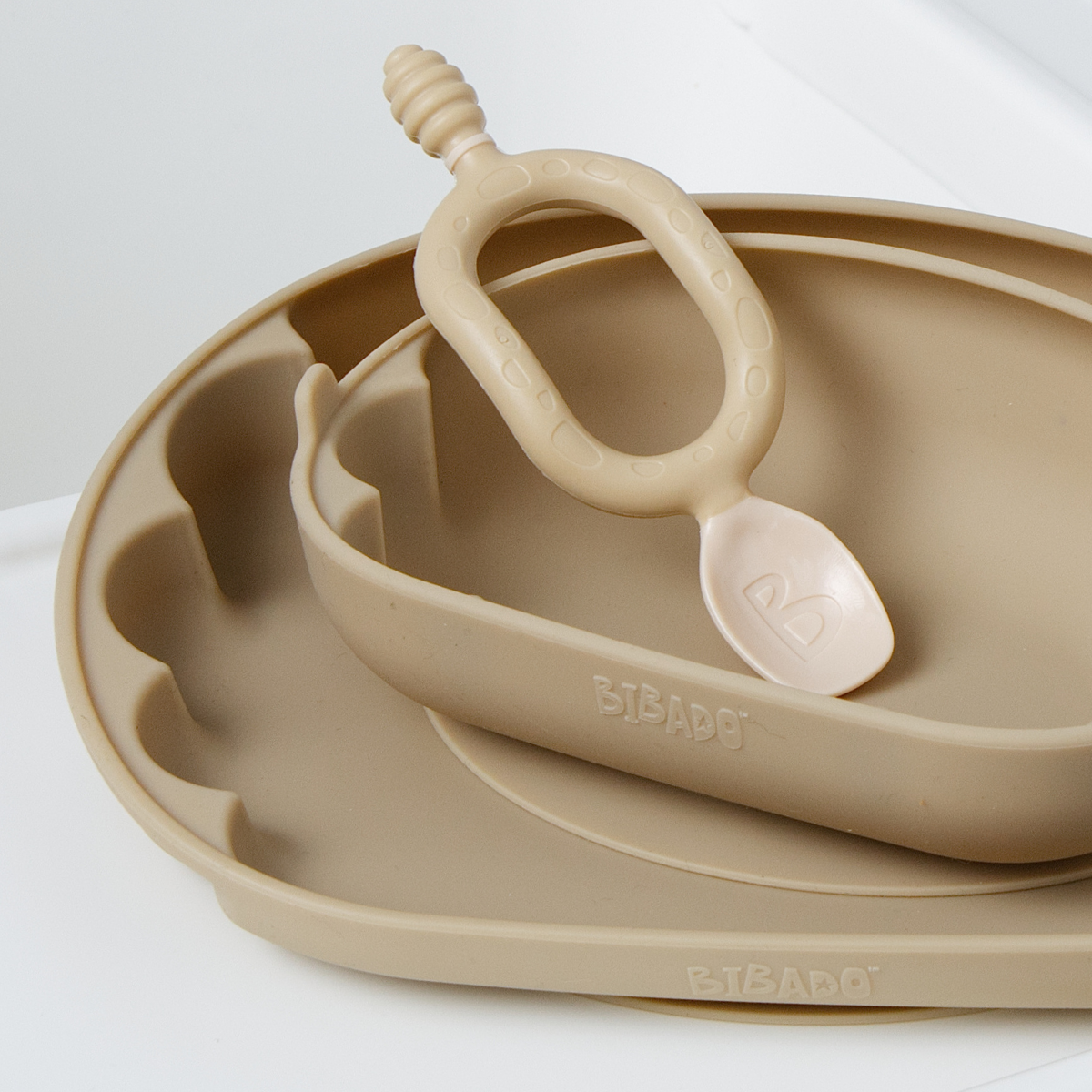 A beige silicone baby feeding set including a scalloped plate, a smaller bowl, and a Dippit spoon shown on a clean white background. The brand name 'Bibado' is embossed on the side of the bowl. - Product image