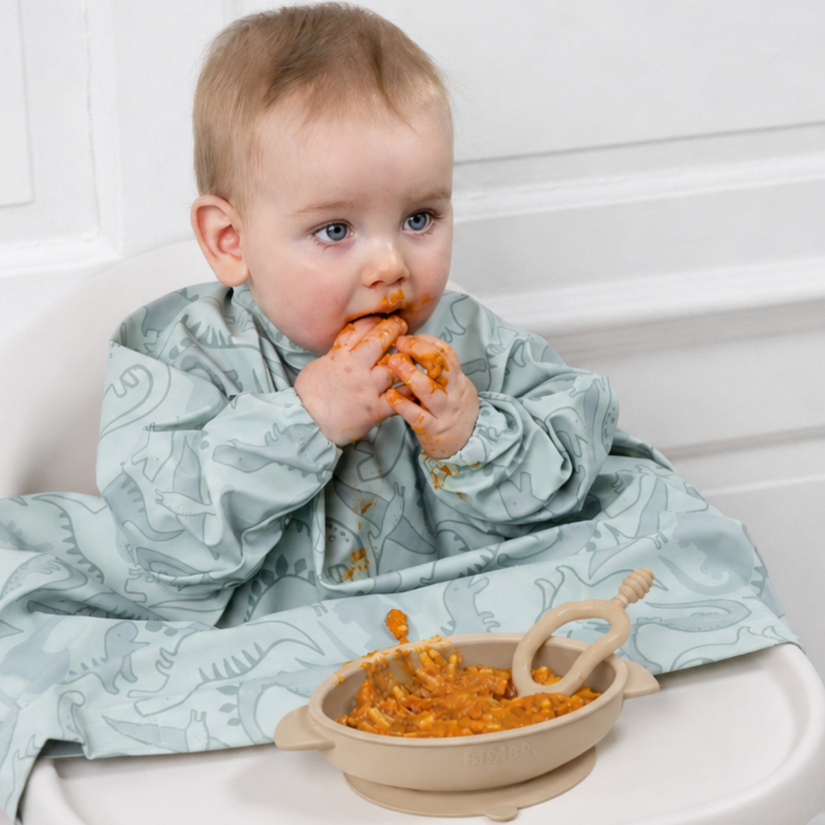 Baby in a light green dinosaur-patterned long-sleeve bib, eating from a beige silicone suction bowl with a matching spoon. - Product image