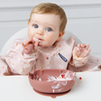 A baby wearing a light pink floral bib sits in a white high chair, eating from a dusty pink silicone bowl. 