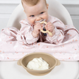 Baby in a pink floral bib, chewing a Dippit spoon, with a beige silicone suction bowl of yogurt.