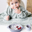 Messy baby with curly hair in a dinosaur bib, looking up, next to a mist silicone suction bowl with yogurt and berries.