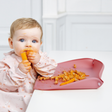 A baby wearing a light pink floral Coverall bib sits in a white high chair, eating a carrot with pasta on a pink feeding mat with the optional sides raised. The baby's face and hands are covered in orange food residue.