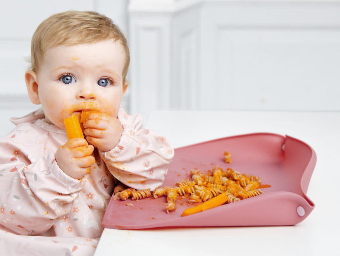 A baby wearing a light pink floral Coverall bib sits in a white high chair, eating a carrot with pasta on a pink feeding mat with the optional sides raised. The baby's face and hands are covered in orange food residue. - image