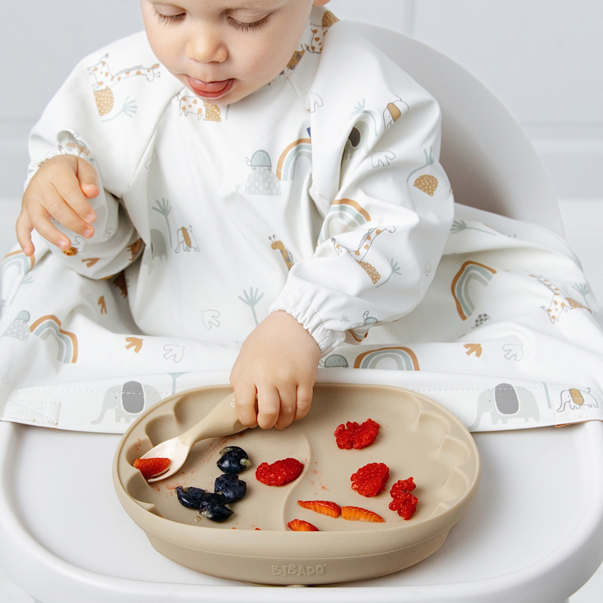 A baby wearing a white long-sleeve bib with a safari animal and rainbow print sits in a high chair, using a beige silicone plate to eat strawberries and blueberries. - Product image