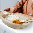 A beige silicone baby feeding plate with scalloped edges, shown on a white high chair. The tray contains white rice, curry, and a small matching silicone spoon.