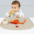 A baby wearing a white Coverall bib sits at a table using a beige silicone baby feeding placemat. On the mat rests a slice of watermelon, strawberries, and a small cream-colored Sippit cup.