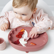 A baby wearing a light pink floral bib sits in a white high chair, looking down at a dusty rose silicone baby plate with a spoon and food pieces. A small matching cup is visible on the left.