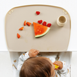 A baby sits at a white table with a beige silicone feeding mat, eating a slice of watermelon and mango. The tray has a small cream-colored cup sat on the right side.
